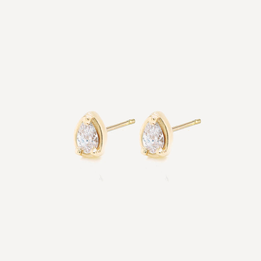 Side view of gold petite pear diamond stud earrings with sparkling pear-cut stones in a polished gold setting on a soft beige background.