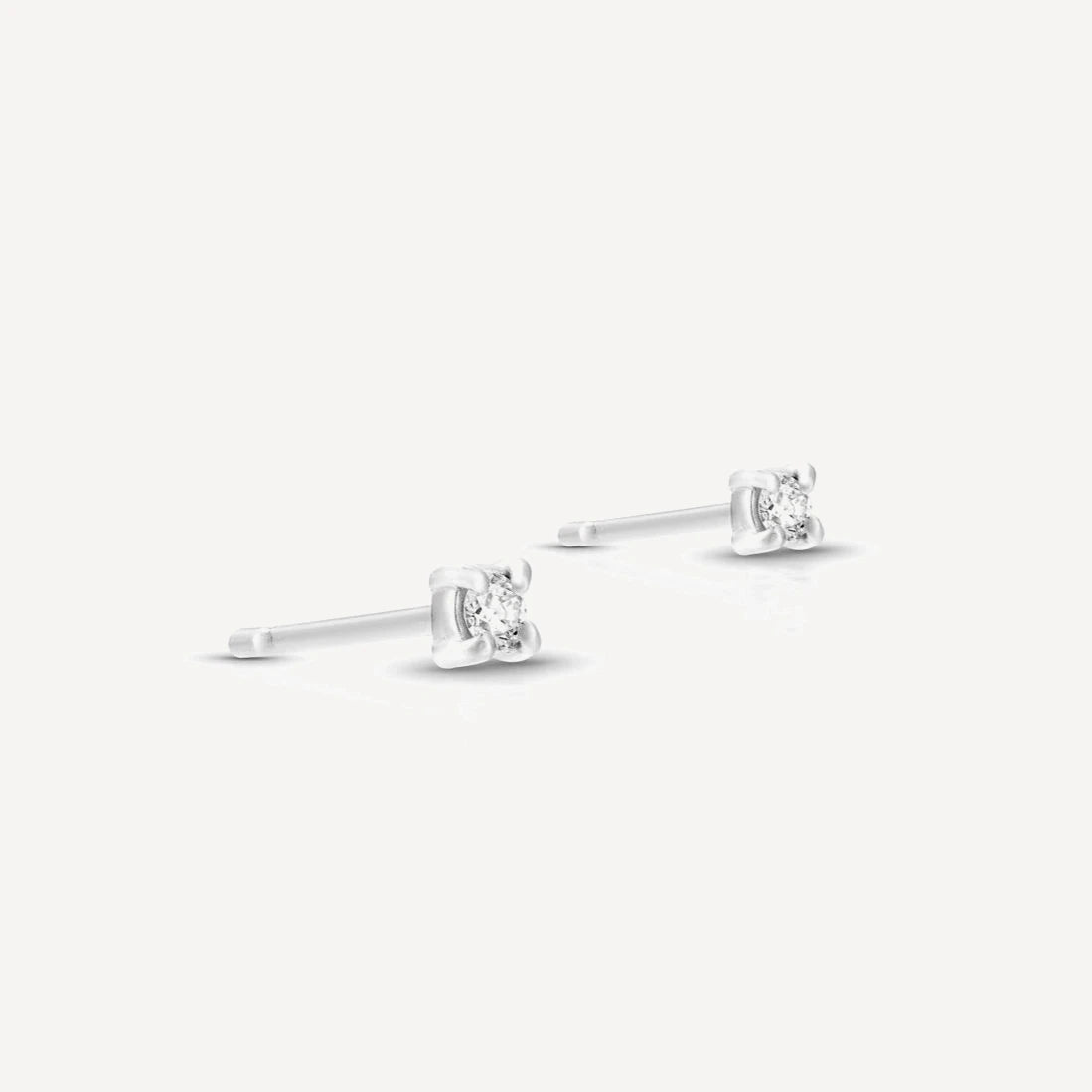 Sterling silver single diamond stud earrings featuring brilliant-cut diamonds in a classic four-prong setting on a soft beige background.