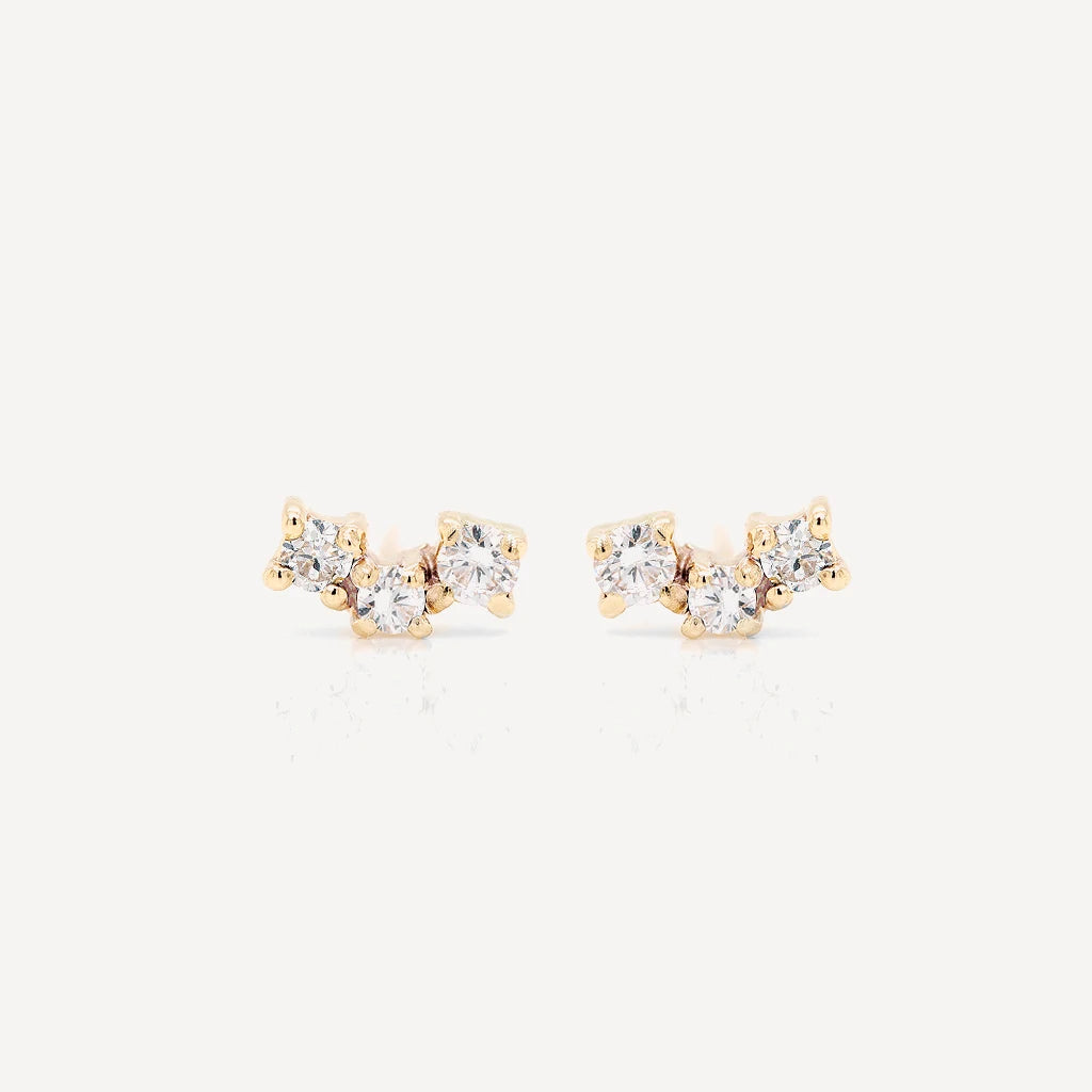 Yellow gold diamond cluster stud earrings featuring three brilliant-cut stones in a delicate clustered design on a soft beige background.