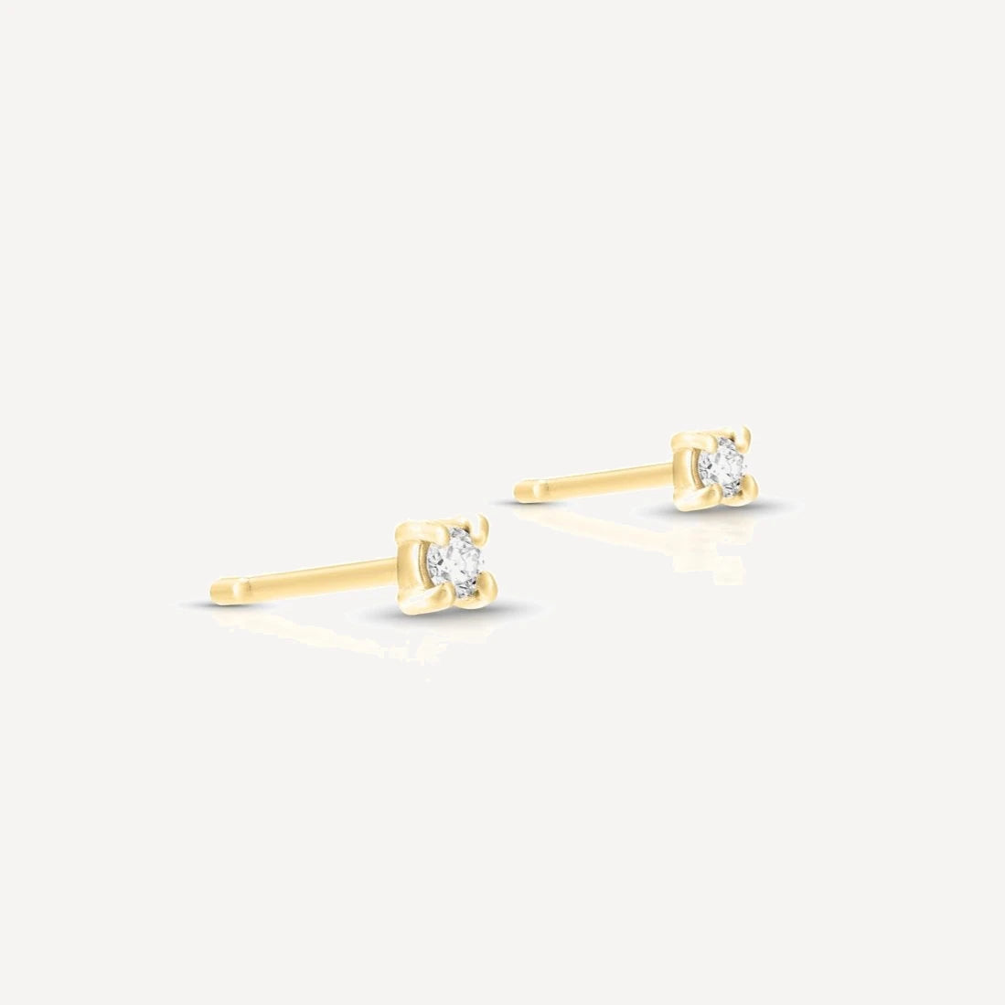 Yellow gold single diamond stud earrings featuring brilliant-cut diamonds in a classic four-prong setting on a soft beige background.